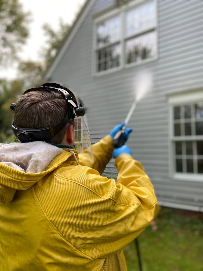 Crafting Captivating Headlines: Your awesome post title goes here Person cleaning house siding with a pressure washer, wearing protective gear in a yellow raincoat.